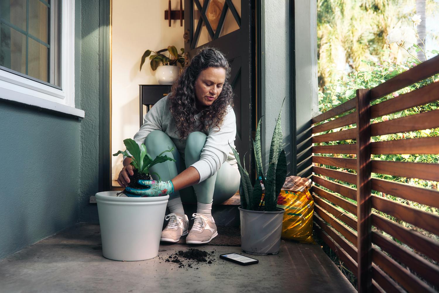 Mulher agachada na varanda de casa cuidando de uma planta, com o celular no chão ao lado.
